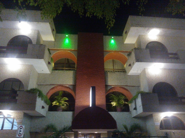 Un edificio de varios pisos con una columna central roja, balcones con plantas en macetas y luces verdes iluminadas.