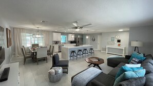 Una sala de estar moderna con un sofá gris, una mesa de centro y un televisor. Incluye una zona de cocina con una isla grande y taburetes de bar.