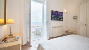 Un cuarto de hotel con una cama, un escritorio con lámpara, un televisor montado en la pared y vista a la playa por una puerta abierta.