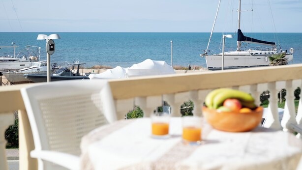 Una mesa con un bol de fruta y dos vasos de zumo de naranja, con vistas a un puerto deportivo con barcos y un cielo azul despejado.