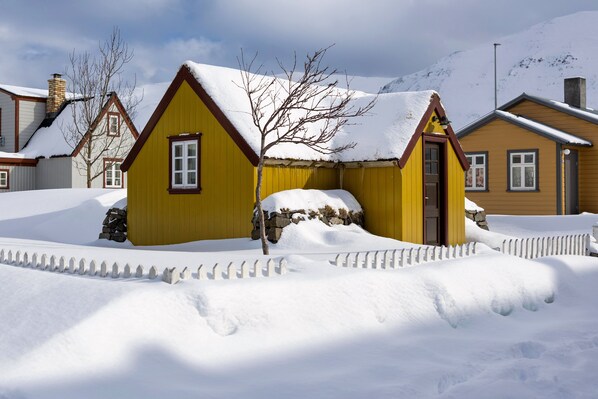 A yellow house with a snow-covered roof and a stone foundation, surrounded by a white picket fence.