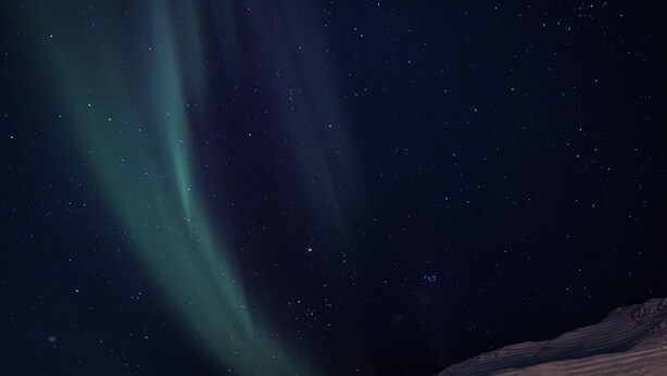 Et snøklædt gade med fodaftryk, der fører hen til huse, under en stjerneklar himmel med nordlys.
