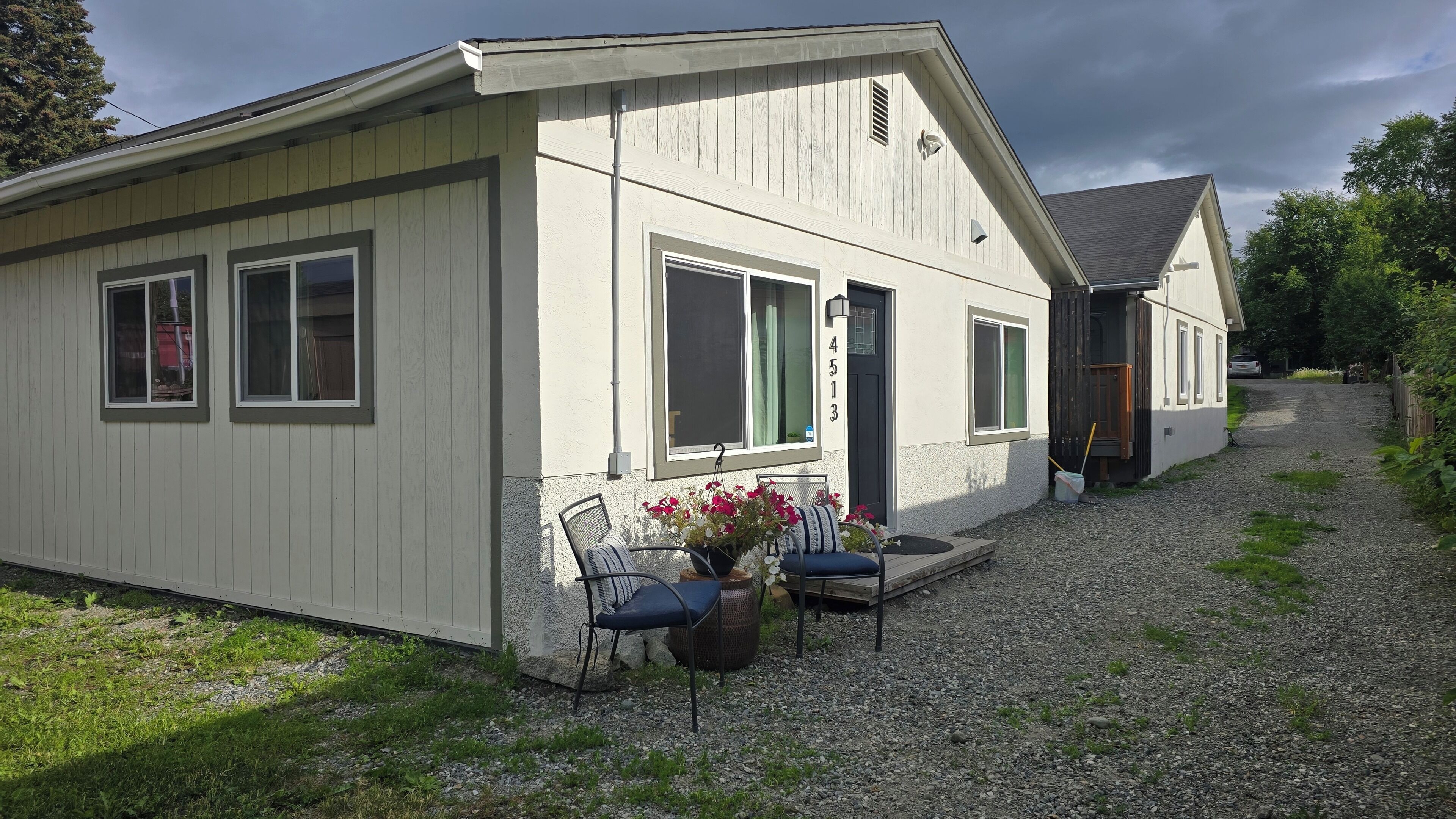 A single-story building with a gravel driveway, a front porch with two chairs and a potted plant, and a visible door number.