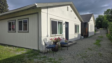 A single-story building with a gravel driveway, a front porch with two chairs and a potted plant, and a visible door number.