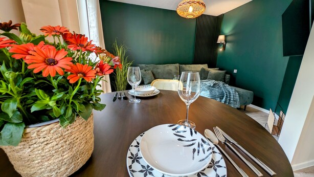 Une table à manger dressée avec des assiettes, des couverts et des verres à vin, entourée d’un panier tressé rempli de fleurs rouges.