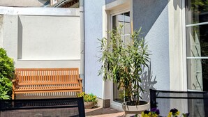 Une terrasse en bois avec un banc et une table, un pot de fleurs, et un bâtiment doté d’un balcon et d’une fenêtre.
