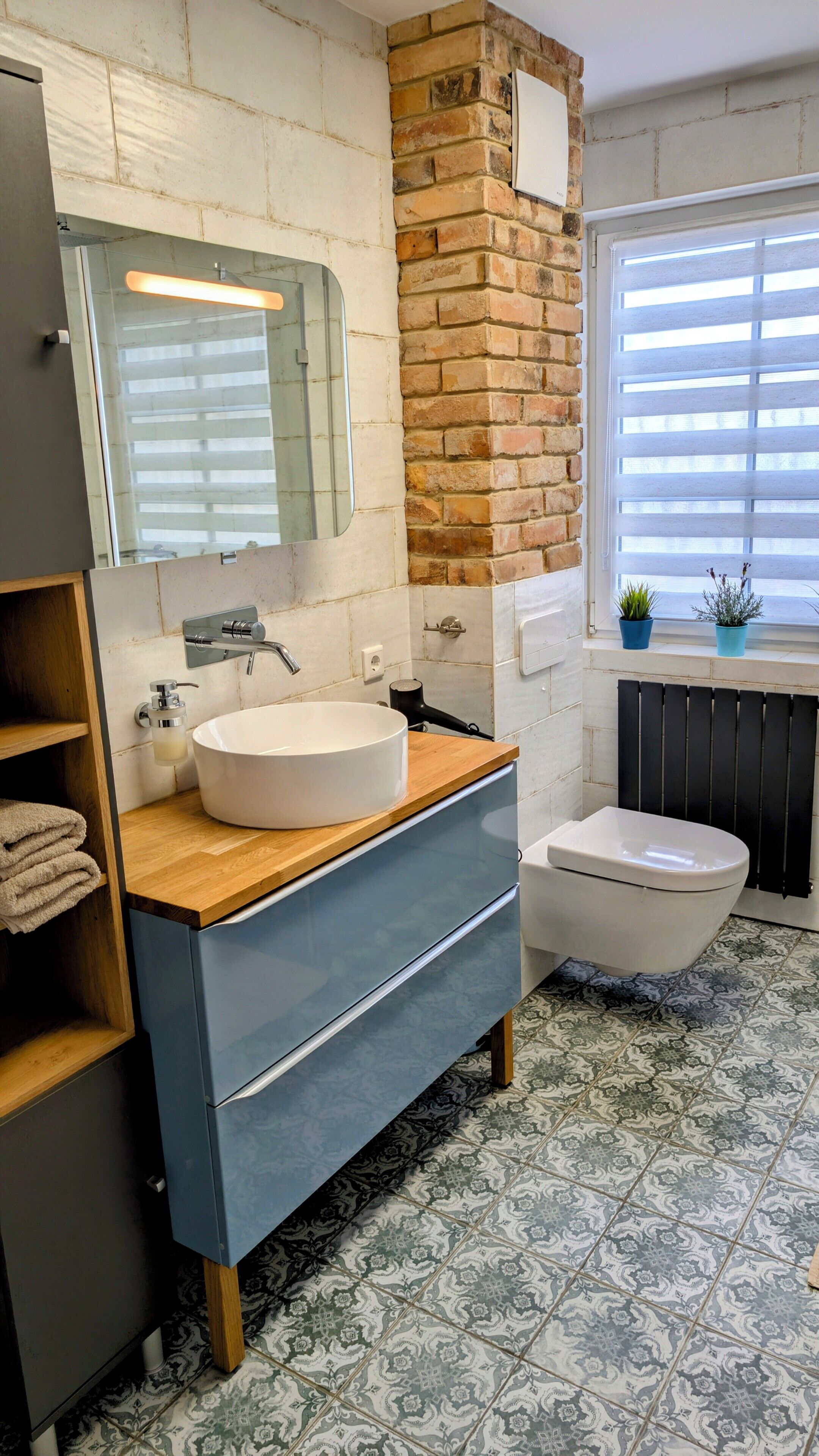 A bathroom with a white sink, a wooden vanity, a mirror, a toilet, and a window with blinds.