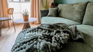 Une chambre avec un mur vert, un lit recouvert d’une couverture grise, une lampe en osier, une chaise, une petite table et une plante en pot.
