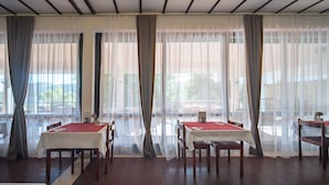 A dining area with tables set for two, large windows with sheer curtains, and a view of the outdoors. - Hotel Issa - Triple Room Sea View 3 (Vis)