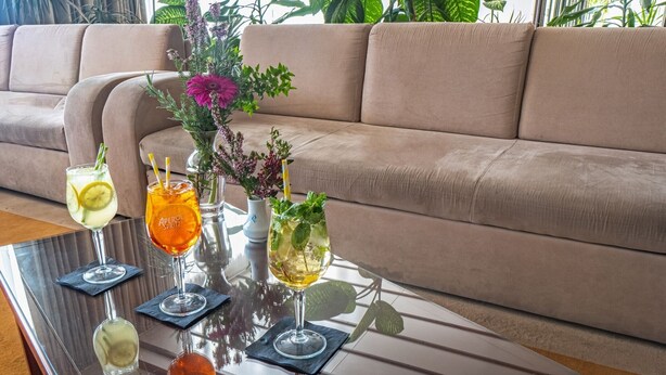 Ein Glastisch mit Getränken und einer Blumenvase in einem Raum mit einem beigefarbenen Sofa.
