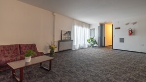 Ein großzügiger Wohnbereich mit einer Couch, einem Couchtisch und einer Topfpflanze. Eine Tür führt in einen anderen Raum, und an der Wand ist ein Feuerlöscher angebracht.
