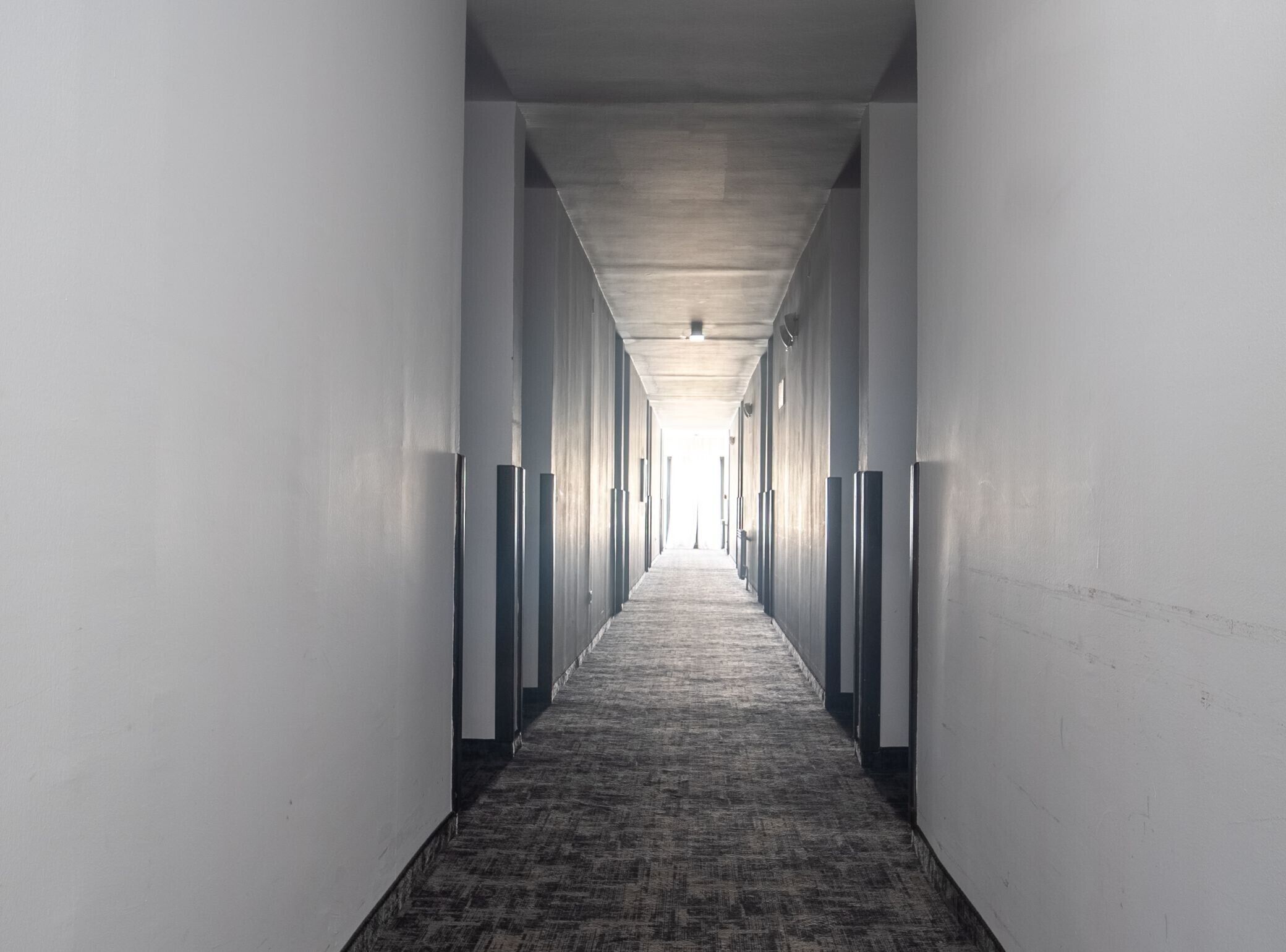 A long, narrow hallway with a carpeted floor and white walls, leading to a bright end.