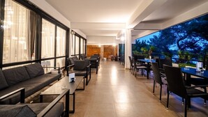 Un área de estar interior moderna con sofás y mesas, ventanas amplias con cortinas, y vistas a árboles a través de un muro de cristal.