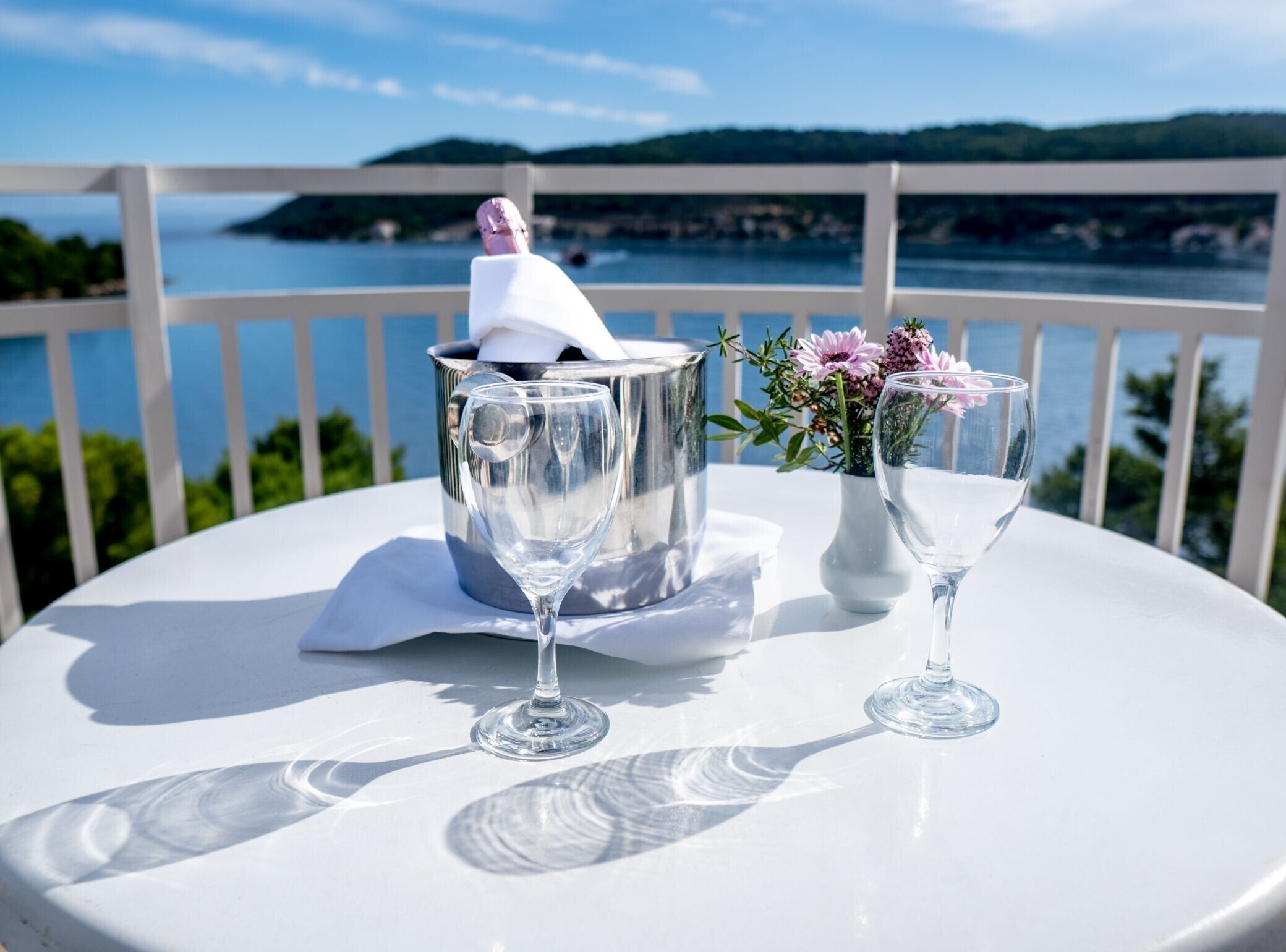 A table with two wine glasses and a flower vase on a balcony overlooking a body of water.