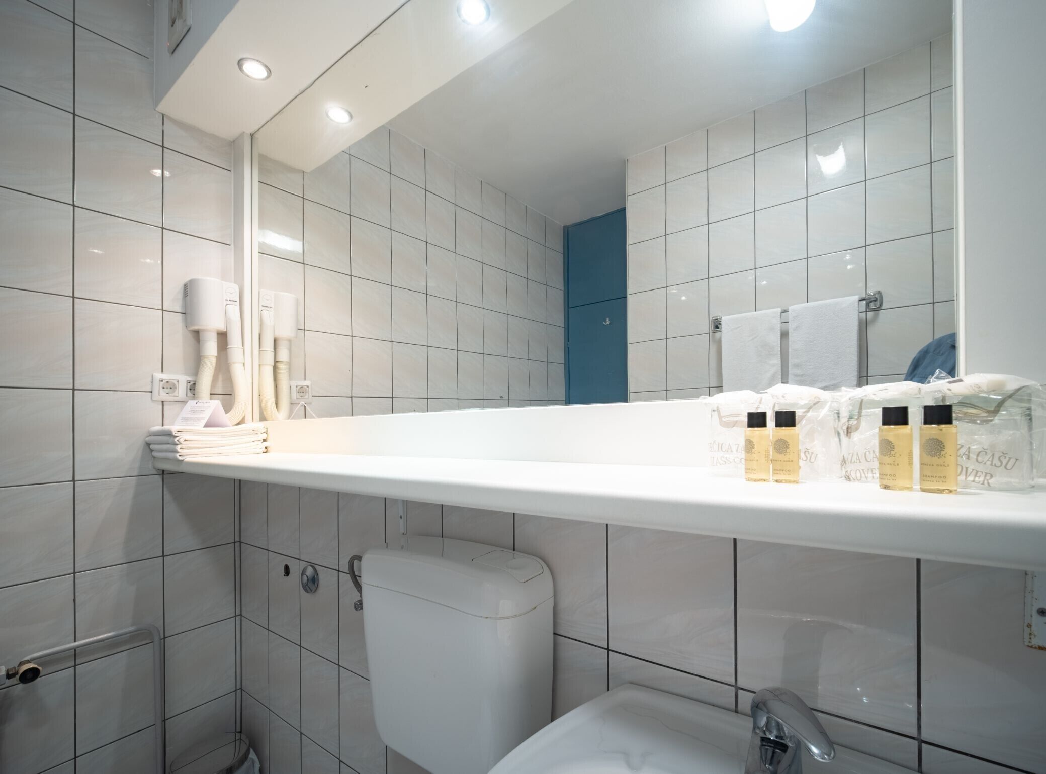 A modern bathroom with a white sink, a large mirror, and a blue door. There are toiletries on the sink and a towel rack with folded towels.