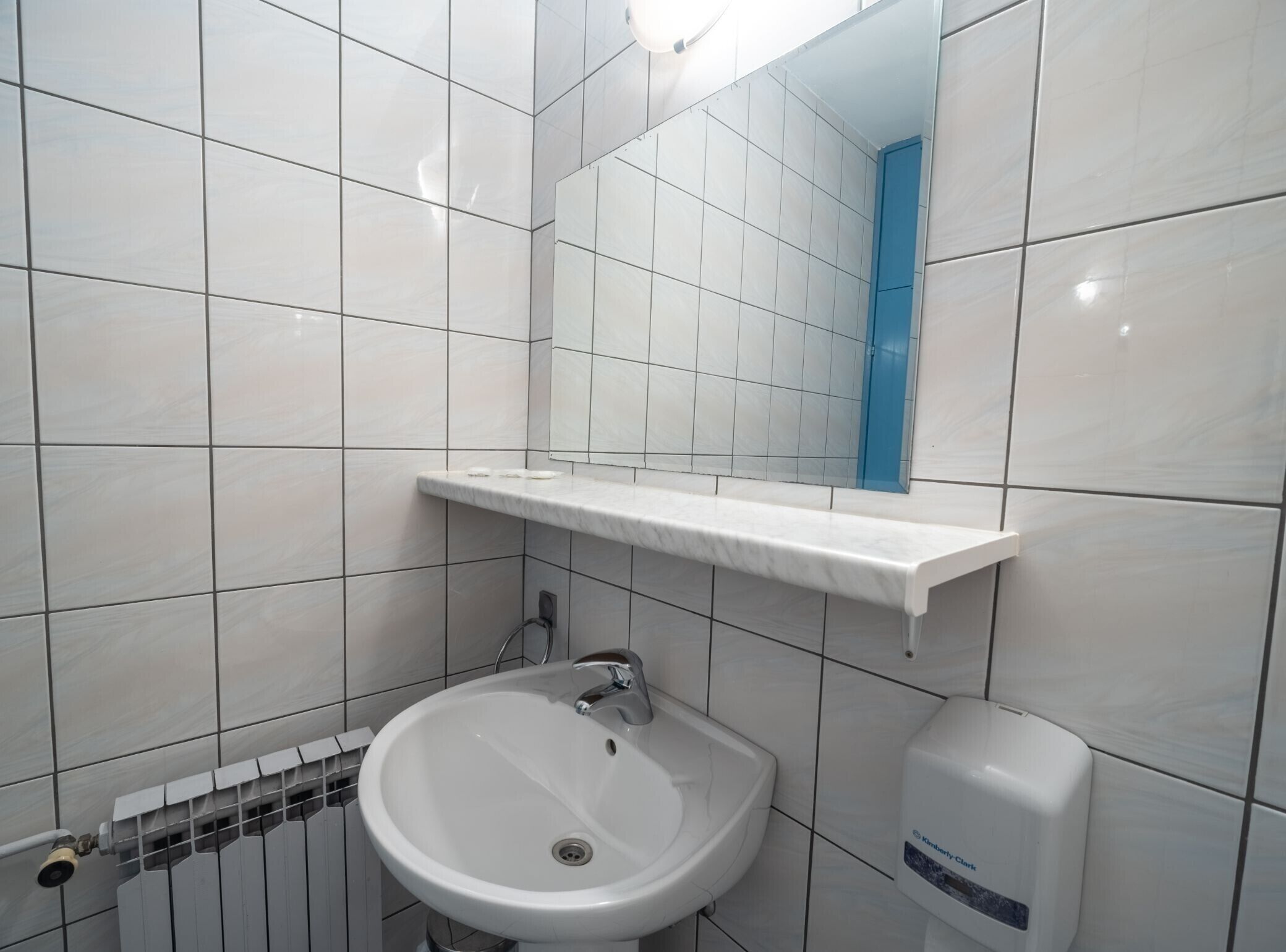 Une salle de bain avec des carreaux blancs, un lavabo blanc, un miroir et une porte bleue.