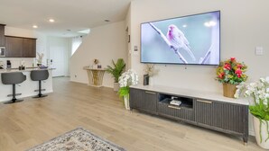 Uma sala de estar moderna com televisão de tela plana, um móvel para centro de entretenimento em madeira escura e uma cozinha ao fundo.