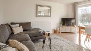 A modern living room with a grey sofa, a coffee table, a TV, and a large window with a view of buildings. - Ty Golau (Pwllheli)