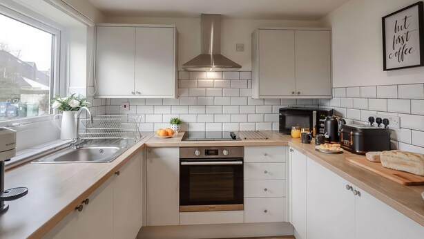 A modern kitchen with white cabinetry, a built-in oven, a coffee machine, and a window providing a view of the outside. - Ty Golau (Pwllheli)