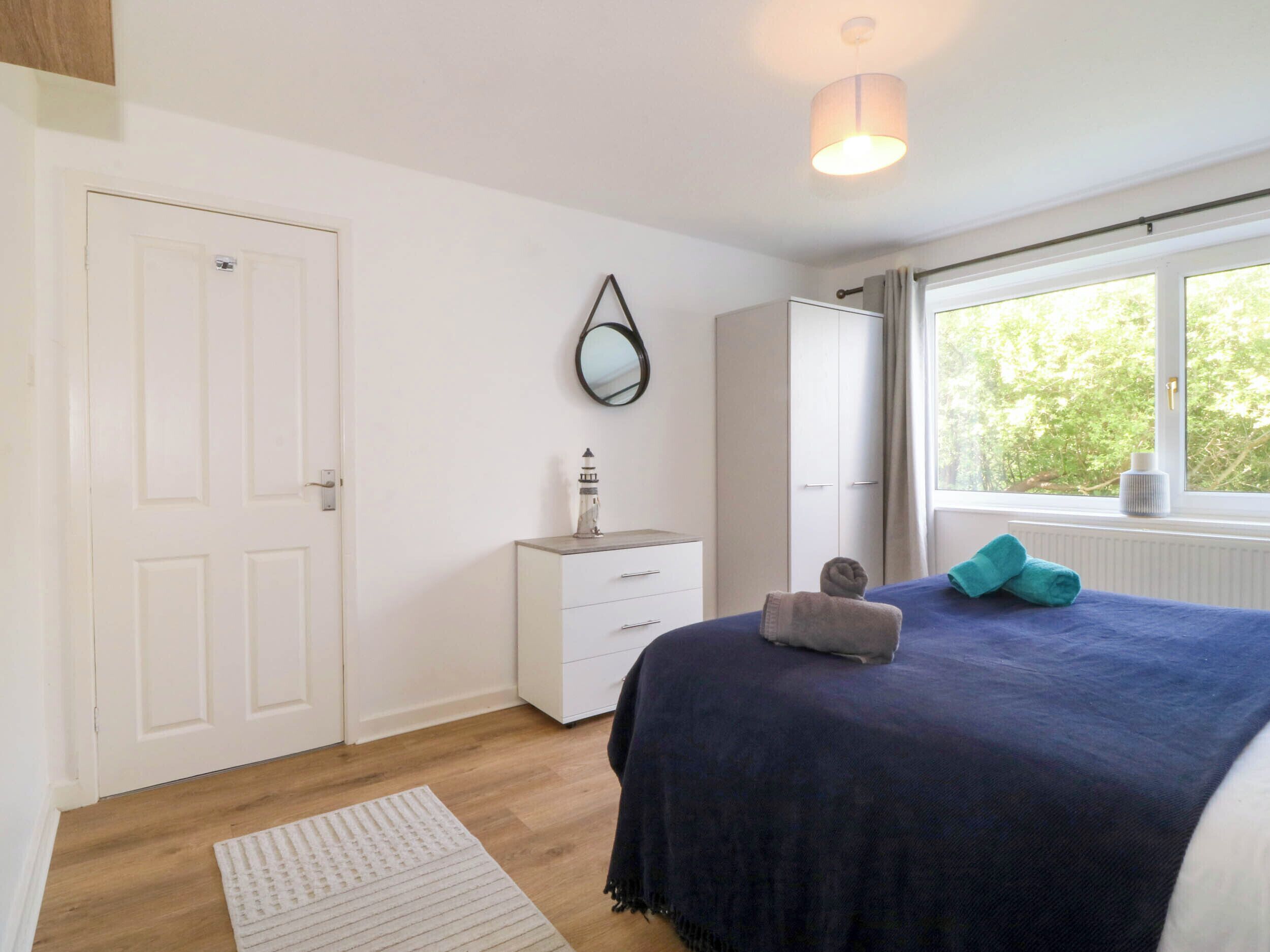 Un dormitorio con cama, cómoda, espejo, ventana y vistas a la vegetación, y una puerta.