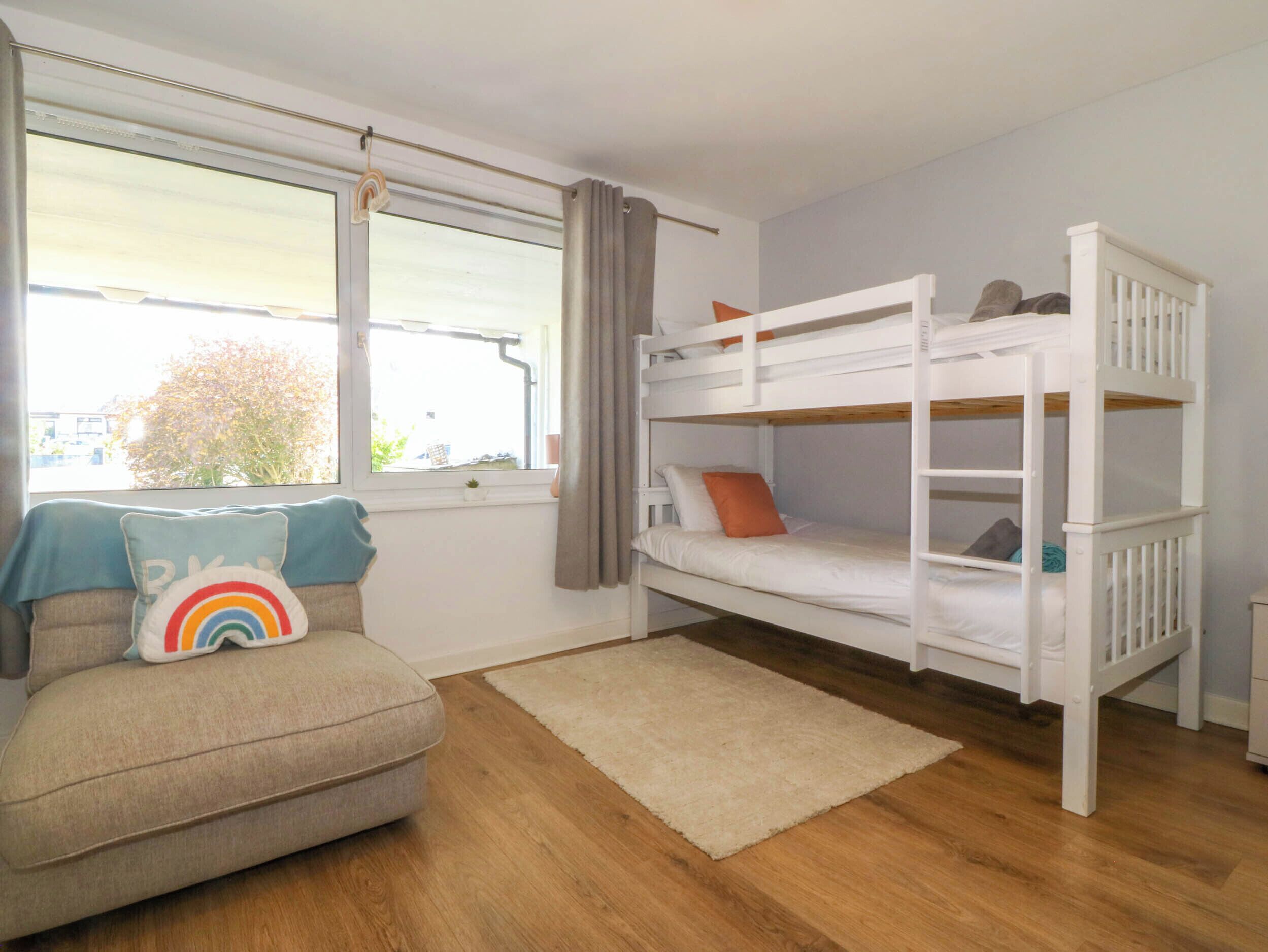 A room with a bunk bed, a sofa with a rainbow pillow, a window with curtains, and a view of a tree outside.