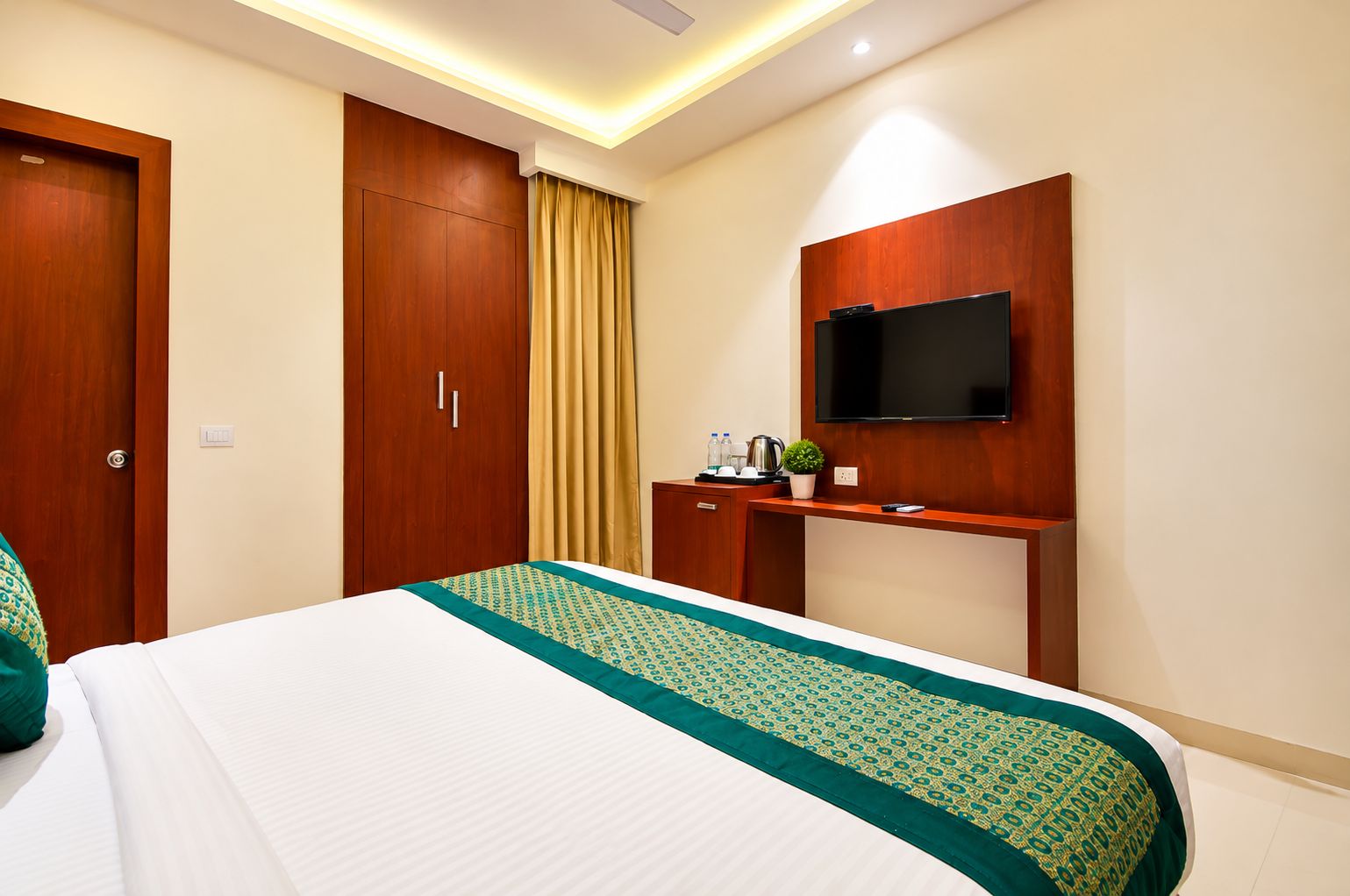 Quarto de hotel com cama, televisão presa a um painel de madeira, secretária com máquina de café e guarda-roupa.