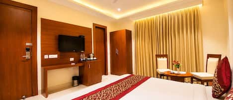 A hotel room with a bed, a desk with a television, a chair, a small table with a vase, and curtains.