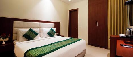 A hotel room with a large bed, a wooden wardrobe, a desk with a kettle, and a nightstand with a vase of flowers.