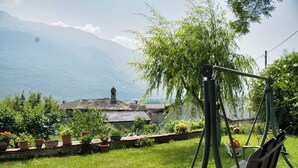 Un jardín con un columpio, plantas en macetas y vistas a una casa y montañas.