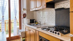 A kitchen with wooden cabinets, a gas stove, a sink, and a window with curtains. - Grigna Mountain View (Narro-indovero)