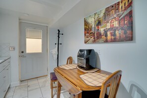 Een eetruimte met een houten tafel, stoelen en een koffieapparaat. Er is een deur met een raam en een schilderij aan de muur.