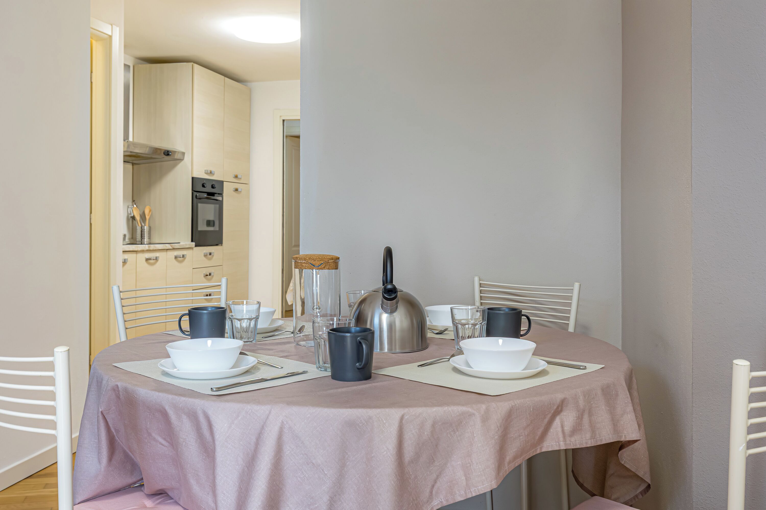 A dining area with a round table set for four, equipped with white plates, bowls, and cutlery. A kettle and a coffee maker are on the table. The room has a neutral color scheme with white walls and a wooden cabinet in the background.
