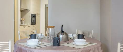 A dining area with a round table set for four, equipped with white plates, bowls, and cutlery. A kettle and a coffee maker are on the table. The room has a neutral color scheme with white walls and a wooden cabinet in the background.