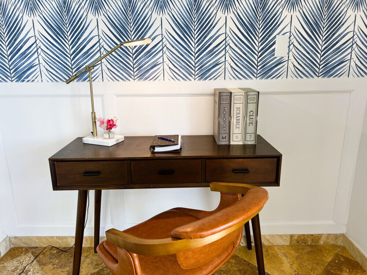 A desk with a leather chair, a lamp, and books on a white wall with a leaf pattern.