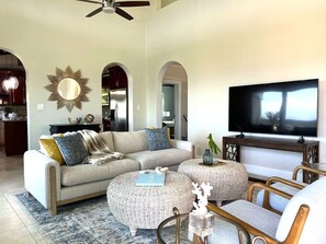 A living room with a sofa, ottomans, a TV, and a ceiling fan.