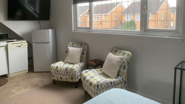 Una habitación pequeña con dos sillones estampados, una cama blanca, un televisor de pantalla plana, un refrigerador y una ventana con vista a casas.