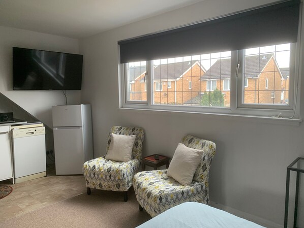 A small room with two patterned armchairs, a white bed, a flat-screen TV, a refrigerator, and a window with a view of houses.
