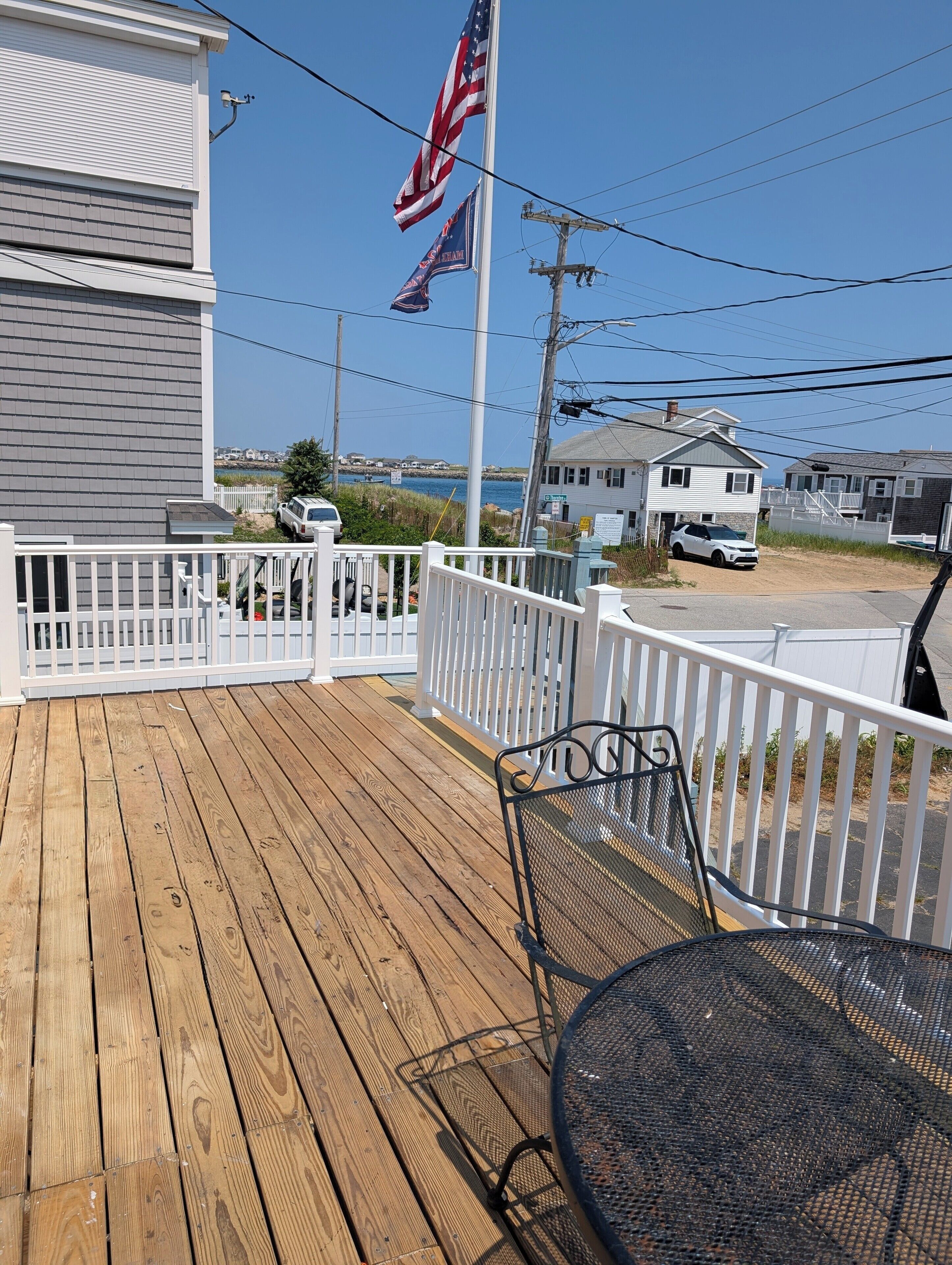 Una terraza de madera con una mesa y sillas de metal, barandal blanco y vista a casas y un asta con banderas.