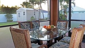 Una mesa de vidrio con sillas, un cuenco de fruta y vistas a una casa y palmeras a través de amplias ventanas.
