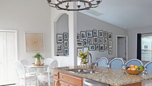 Una cocina moderna con una isla central, electrodomésticos de acero inoxidable y una araña. Hay cuadros enmarcados en la pared, un comedor con sillas blancas y un ventanal grande.