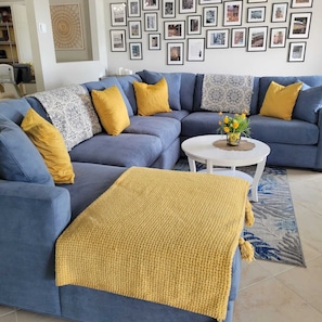 Un salon avec un canapé d’angle bleu, des coussins décoratifs jaunes, une table basse blanche et un tapis à motifs.