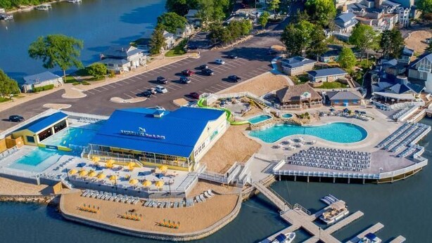 An aerial view of a waterfront hotel with a large pool, a separate building with a blue roof, and a dock area.