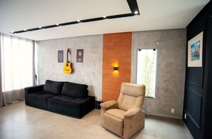 A modern living room with a black sofa, a beige armchair, a guitar, and a brick accent wall. - Casa pet Friendly c Piscina Churrasqueira e Wi-fi (Saquarema)