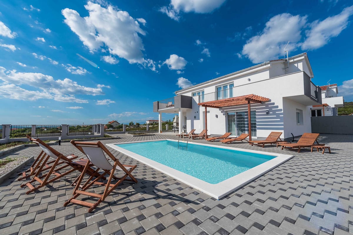 A modern two-story house with a swimming pool, outdoor seating, and a clear blue sky.