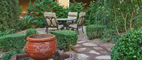 An outdoor patio with a round table and chairs, surrounded by neatly trimmed hedges and a large terracotta pot.
