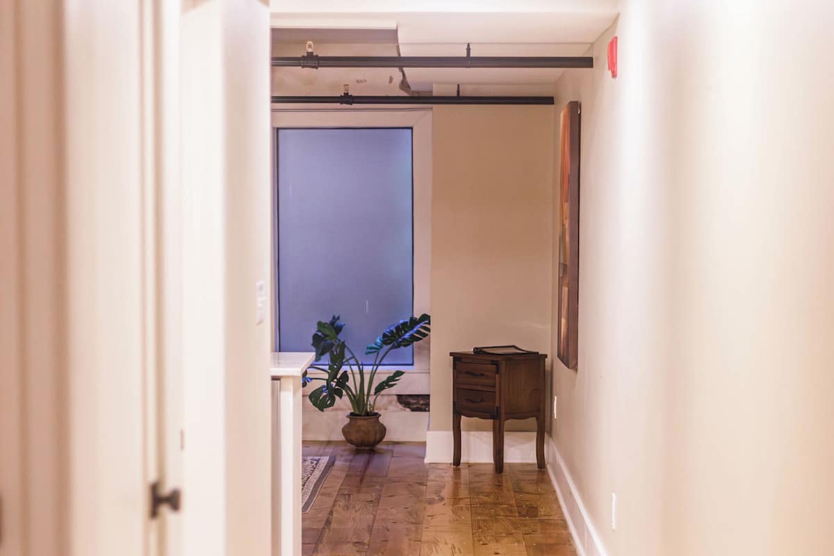 Een hal met houten vloeren, een witte deur, een kleine tafel met een plant en een lijst met een afbeelding aan de muur.