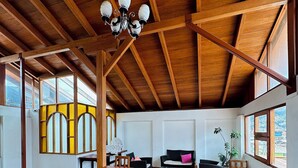 Amplia habitación con vigas de madera, un candelabro, una zona de comedor con mesa y sillas, un sofá y grandes ventanales con vistas.