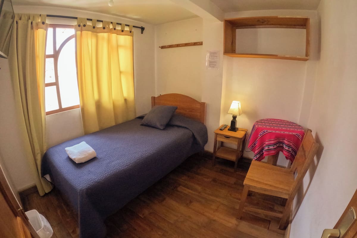 A small, cozy room with a single bed, a nightstand with a lamp, and a small table with a red cloth.