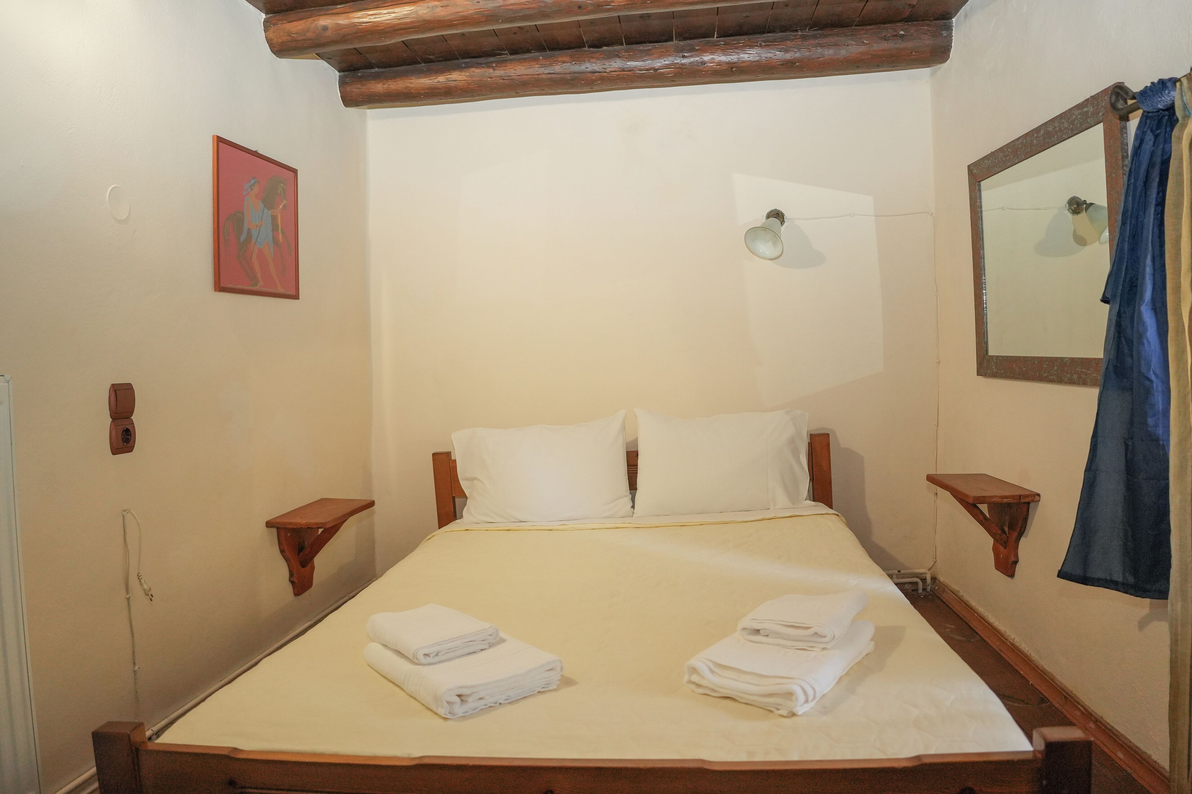 Cama bem arrumada com lençóis e toalhas brancos, cabeceira de madeira, mesas de cabeceira, uma luminária fixada na parede e um espelho.
