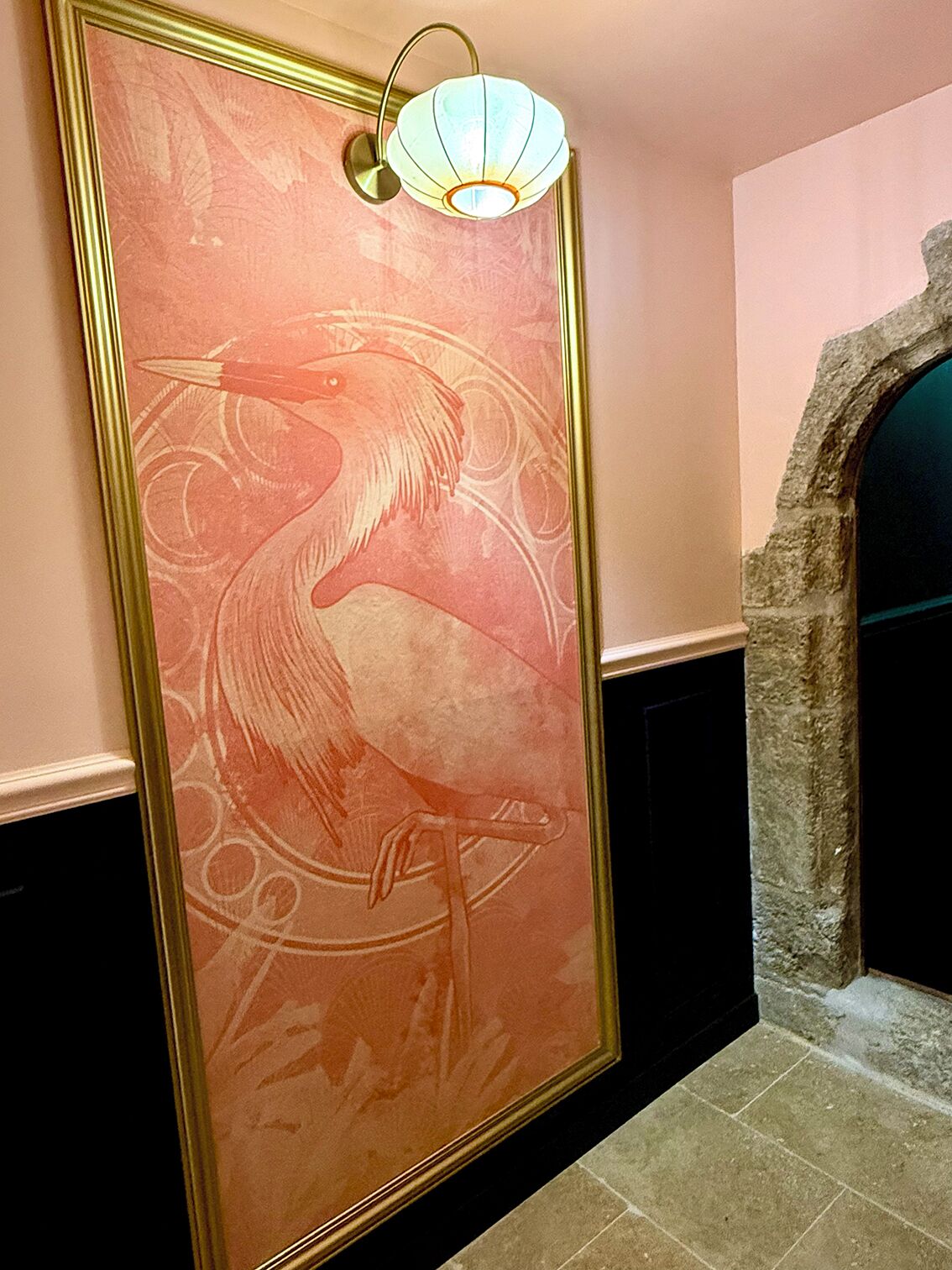 A wall with a large, ornate painting of a bird, a stone archway, and a wall-mounted light fixture.